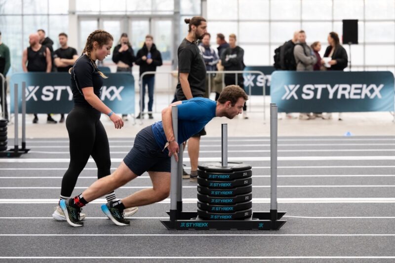 Man in een blauw shirt voert een zware sled push uit op een indoor baan tijdens een Styrekx evenement, nauwlettend geobserveerd door een scheidsrechter.