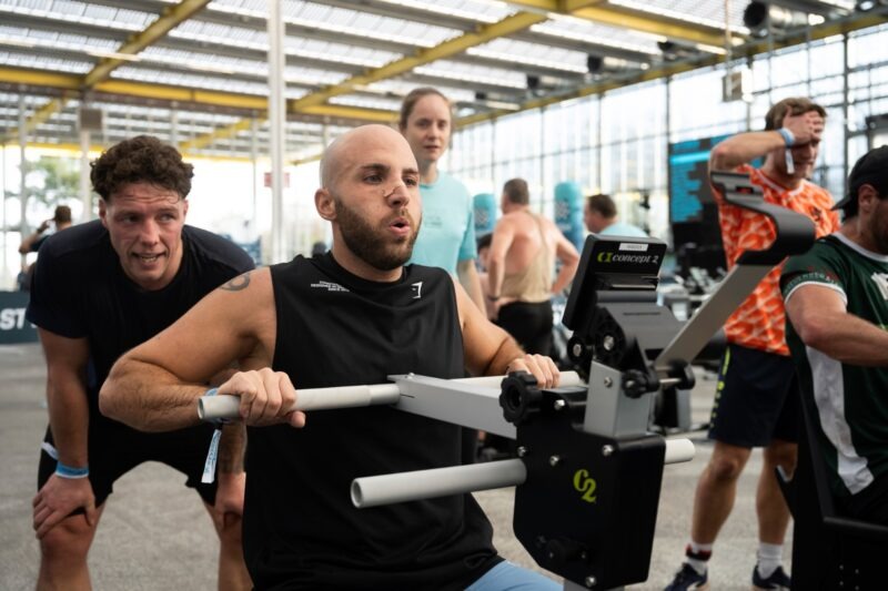 Een mannelijke atleet levert een zware inspanning tijdens de 'Strength Erg' oefening op een Concept2 apparaat, terwijl een medesporter of coach voorovergebogen naast hem staat om hem fanatiek aan te moedigen.