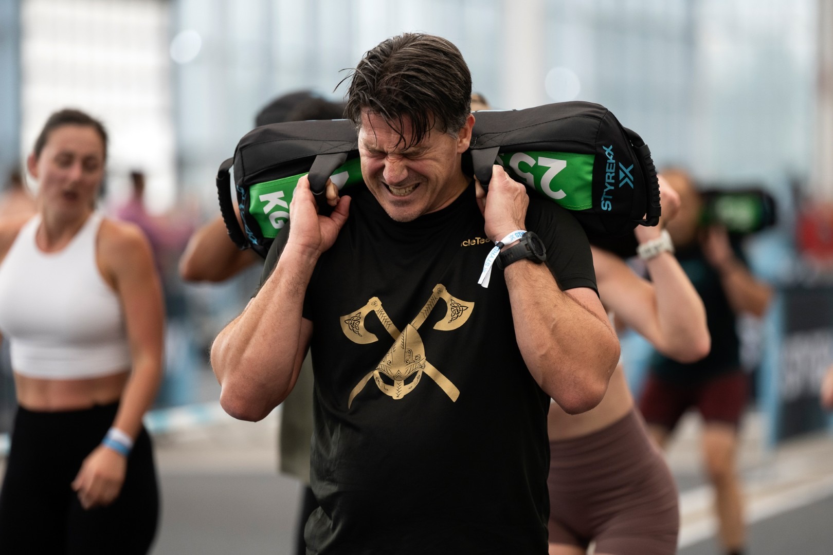 Een geconcentreerde man voert sandbag lunges uit tijdens een Styrekx-fitnessevenement. Hij draagt een zwarte en groene Styrekx-zandzak van 25 kg op zijn schouders en een zwart Styrekx-sportshirt. Hij bevindt zich in een diepe lunge-positie op een genummerde, gemarkeerde grijze indoor baan in een grote, lichte sporthal met industriële uitstraling. Op de achtergrond kijken andere atleten toe en rusten uit, en er is een groot Styrekx-spandoek zichtbaar.