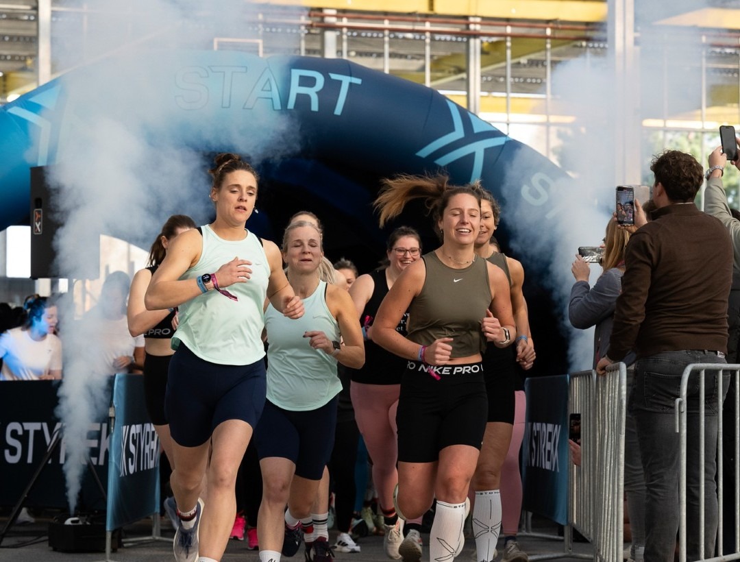 Een groep vrouwelijke sporters rent lachend en vol energie onder een grote blauwe opblaasbare 'START' boog vandaan door een wolk van witte rook aan het begin van het Styrekx fitnessevenement.